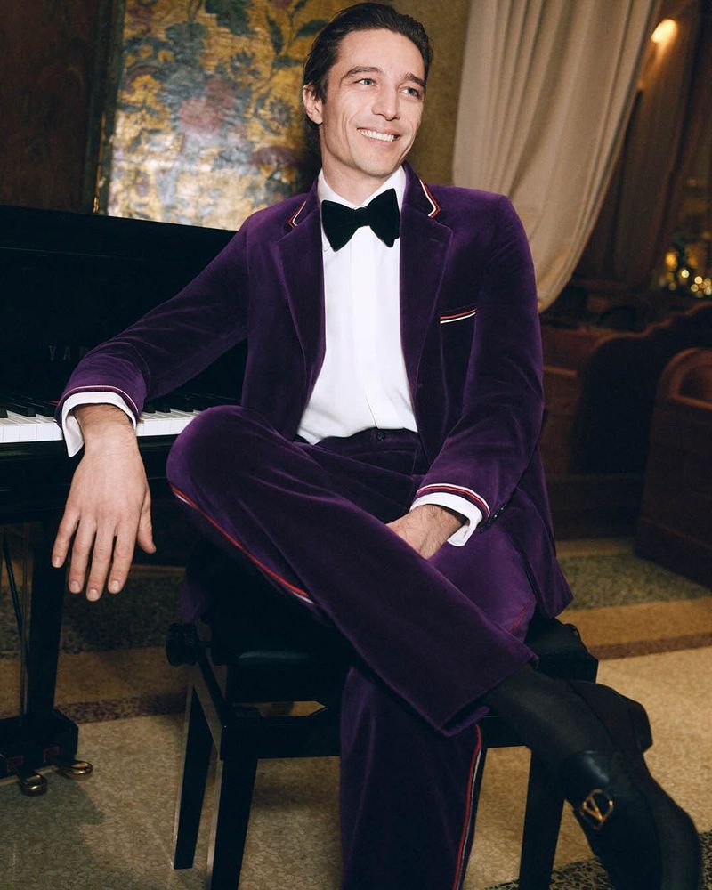 Smiling man in a velvet suit sits near a piano in a hotel lobby with ambient lighting and purple and gold tones. Black boots visible.