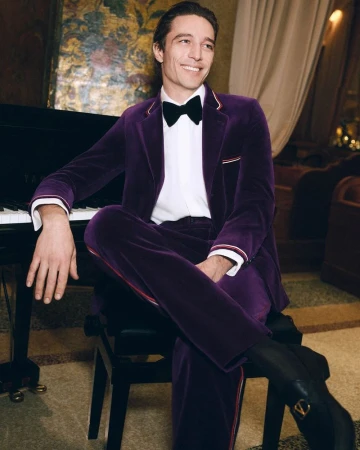 Smiling man in a velvet suit sits near a piano in a hotel lobby with ambient lighting and purple and gold tones. Black boots visible.