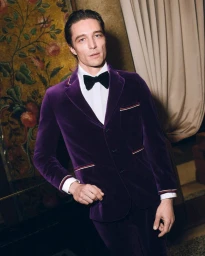 Man wearing a purple velvet suit jacket and black bow tie standing against an ornate interior background