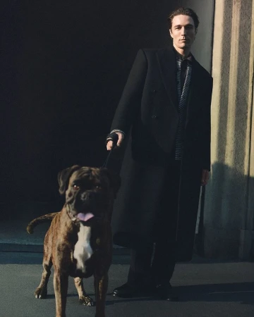 Man wearing a black coat standing on a street holding a leash with a brindle boxer dog in the foreground.