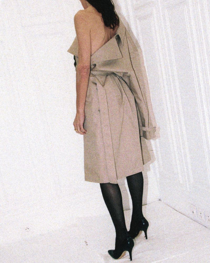 Model wearing a tan trench coat off the shoulder with black tights and heels against a white paneled wall