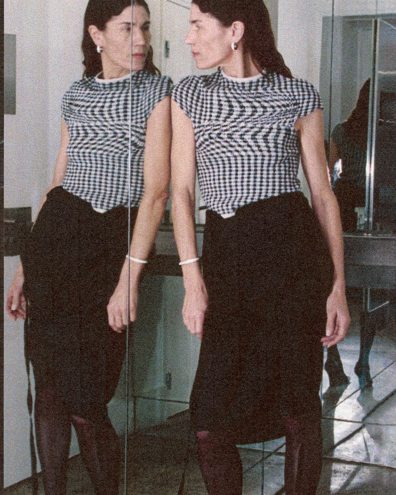 Woman in checkered top and black skirt poses near mirror, showcasing fashion style in an indoor setting with natural lighting.