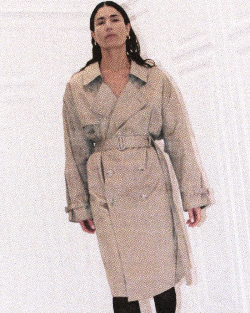 A woman with long dark hair in a light brown trench coat standing against a neutral backdrop, exuding understated fashion.