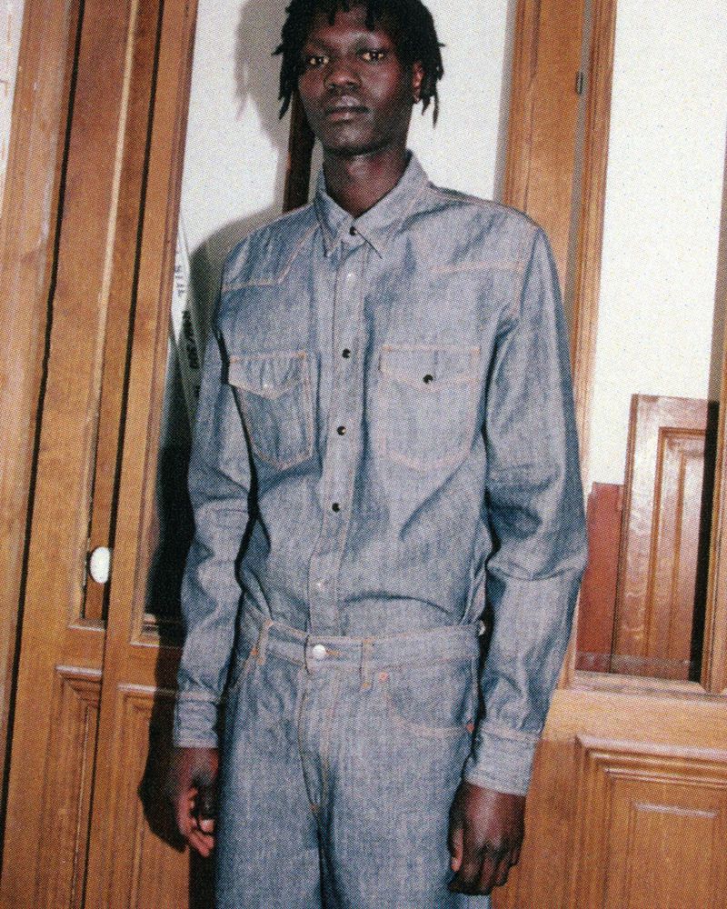 An adult African male stands indoors wearing a denim shirt and pants, with a wooden door in the background, in medium shot.