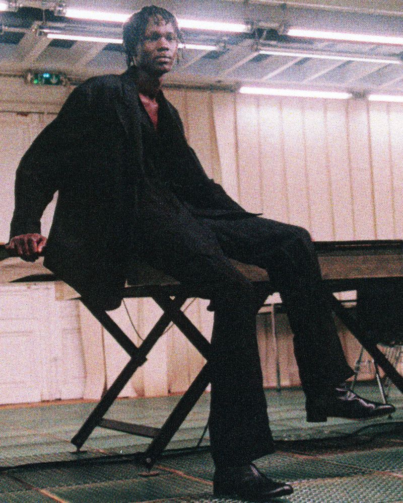 African American man in black suit posing indoors. Male model seated on table in dimly lit room, wearing dark suit and shiny black dress shoes.