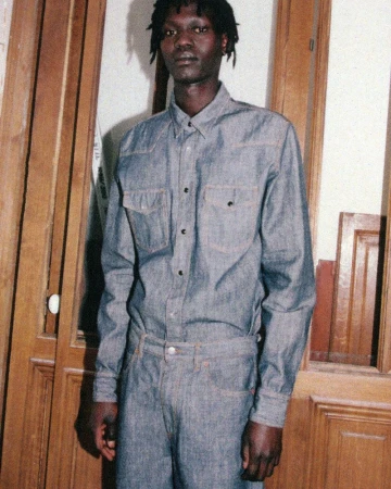 Man wearing a blue denim shirt and matching denim trousers posing inside a room with wooden doors.