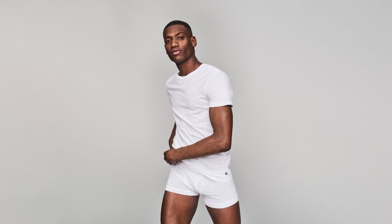A dark-skinned man poses in a white t-shirt and boxer briefs in a full shot against a neutral grey backdrop in a studio environment.