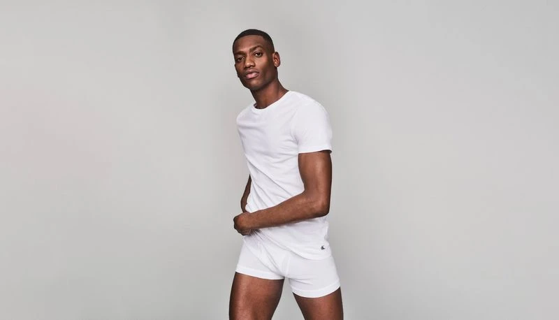 A dark-skinned man poses in a white t-shirt and boxer briefs in a full shot against a neutral grey backdrop in a studio environment.