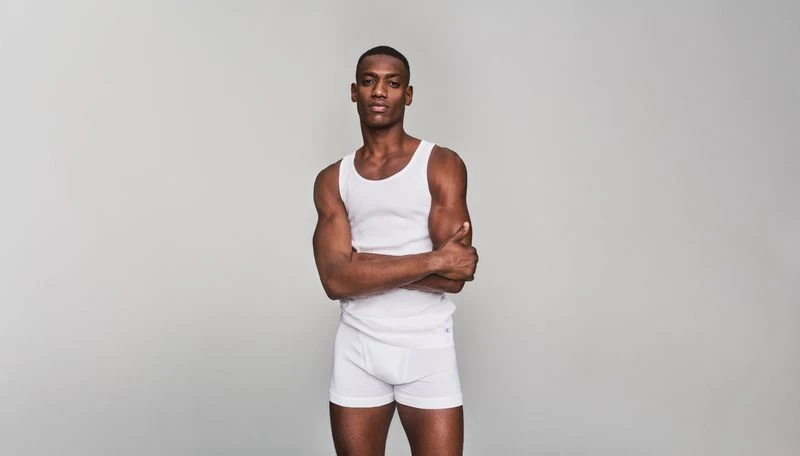 Full-length portrait of a muscular Black man in a white tank top and underwear standing with arms crossed against a gray background.
