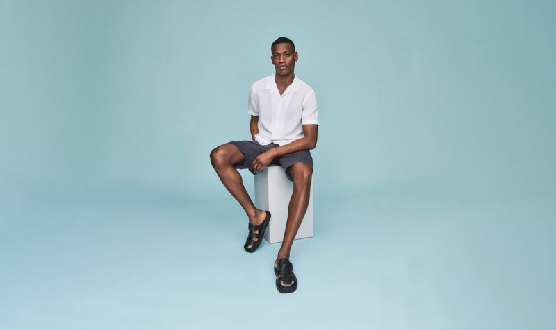 Young man sits on a white block wearing a white short-sleeve shirt, dark shorts, and black sandals against a light blue background.