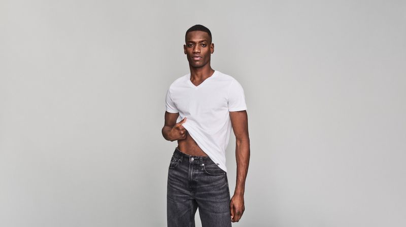 A fit dark-skinned adult male model poses in a white V-neck t-shirt and black jeans, standing in front of a neutral gray background.