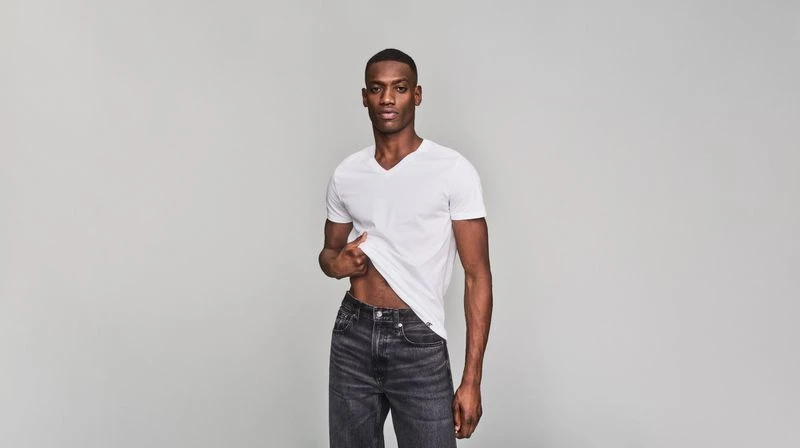 A fit dark-skinned adult male model poses in a white V-neck t-shirt and black jeans, standing in front of a neutral gray background.