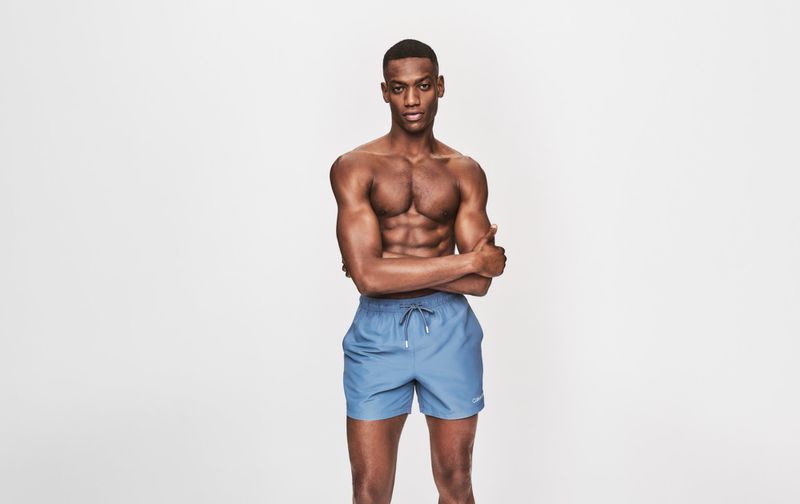 Athletic African American man in blue swim trunks poses shirtless in a studio with a neutral expression, arms crossed and looking forward.