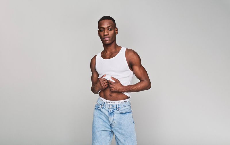 Portrait of a fit Black man in a white tank top and light-wash jeans, striking a confident pose in a neutral photo studio.