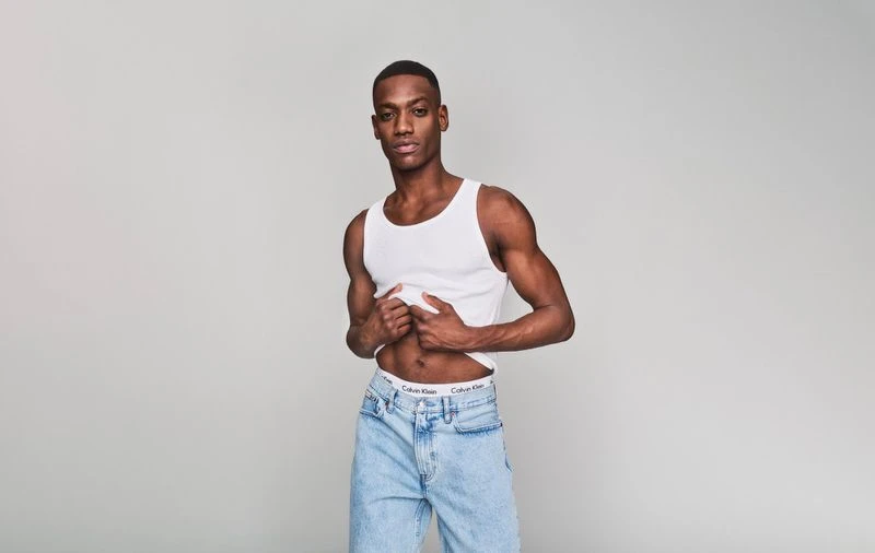 Portrait of a fit Black man in a white tank top and light-wash jeans, striking a confident pose in a neutral photo studio.