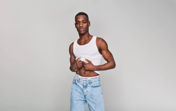 Portrait of a fit Black man in a white tank top and light-wash jeans, striking a confident pose in a neutral photo studio.