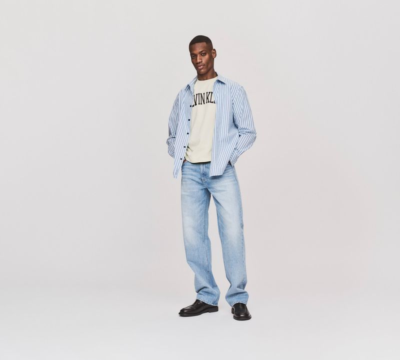 Man wearing a striped shirt over a graphic t-shirt and light blue jeans against a grey seamless background