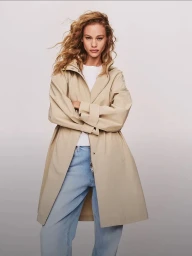 Model with long blonde curly hair wearing a beige coat over blue jeans against a grey seamless background