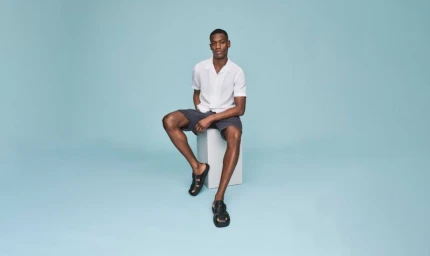 Man seated on a white block wearing a white short sleeve shirt, dark shorts, and black sandals against a solid light blue background.