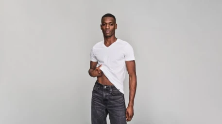 Man wearing a white v neck t shirt and dark jeans posing against a grey studio backdrop