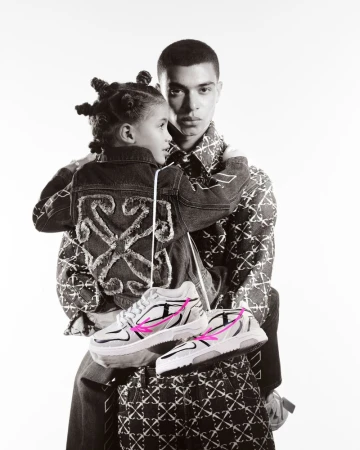 A black and white photograph features an adult and child in patterned jackets holding a pair of athletic shoes with pink accents in a studio setting.