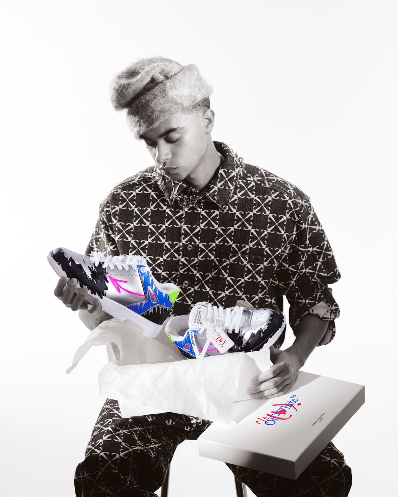 Man wearing a hat and patterned clothing sitting with sneakers in a box, holding one sneaker. White background in an indoor studio.