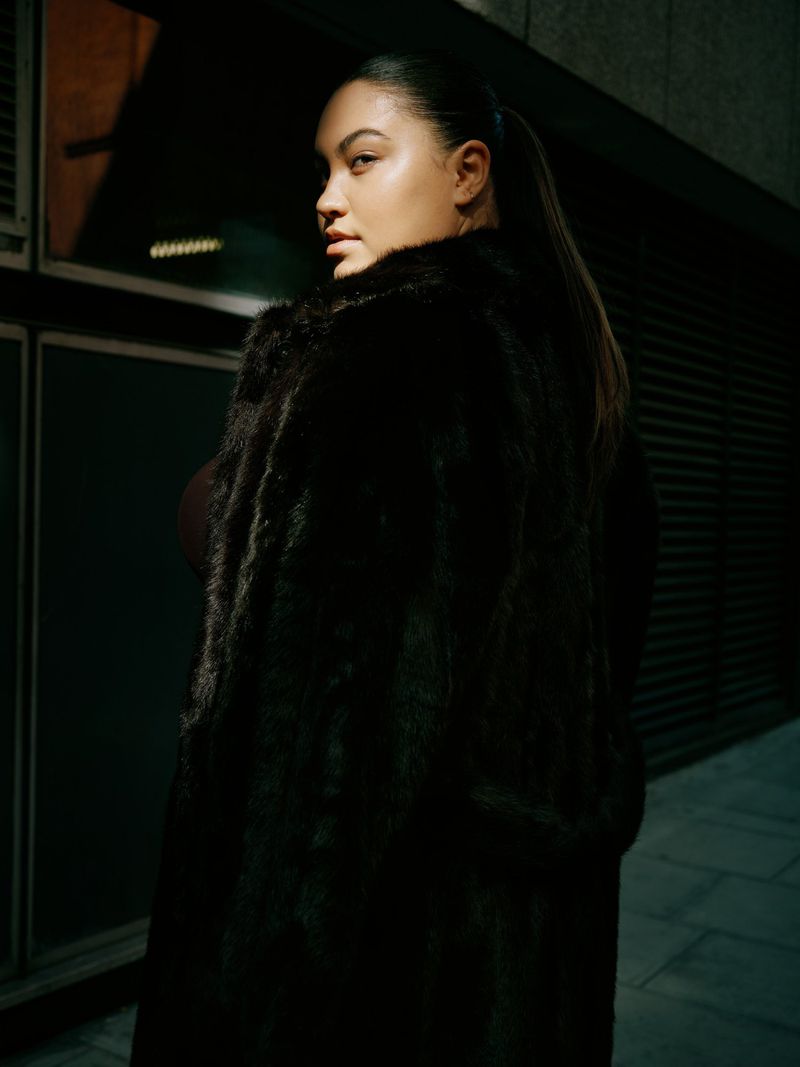 Woman wearing a dark faux fur coat looking back over her shoulder in a dimly lit urban setting at night