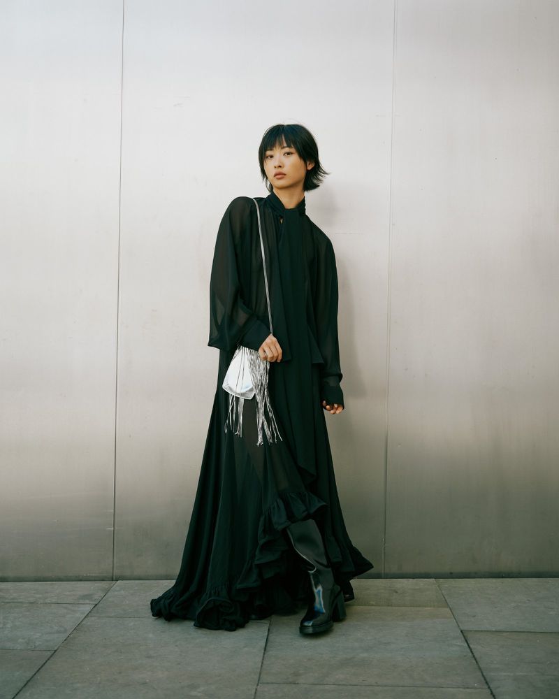 Young woman in a black dress with a silver fringe purse stands against a neutral wall looking at the camera in a fashion editorial style.