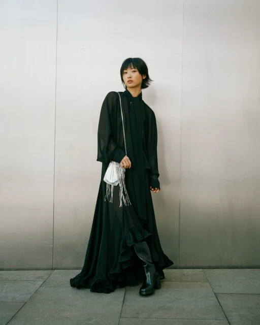 Young woman in a black dress with a silver fringe purse stands against a neutral wall looking at the camera in a fashion editorial style.