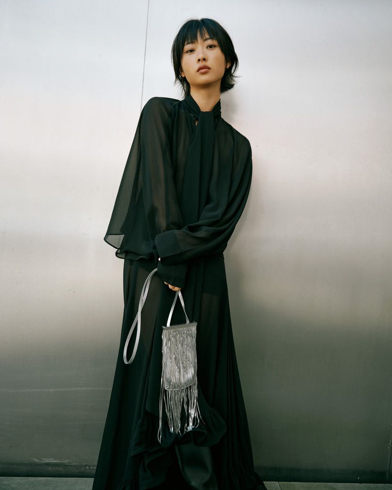A woman in a long black dress and a silver purse stands against a textured metal wall. Fashion editorial portrait with studio lighting.