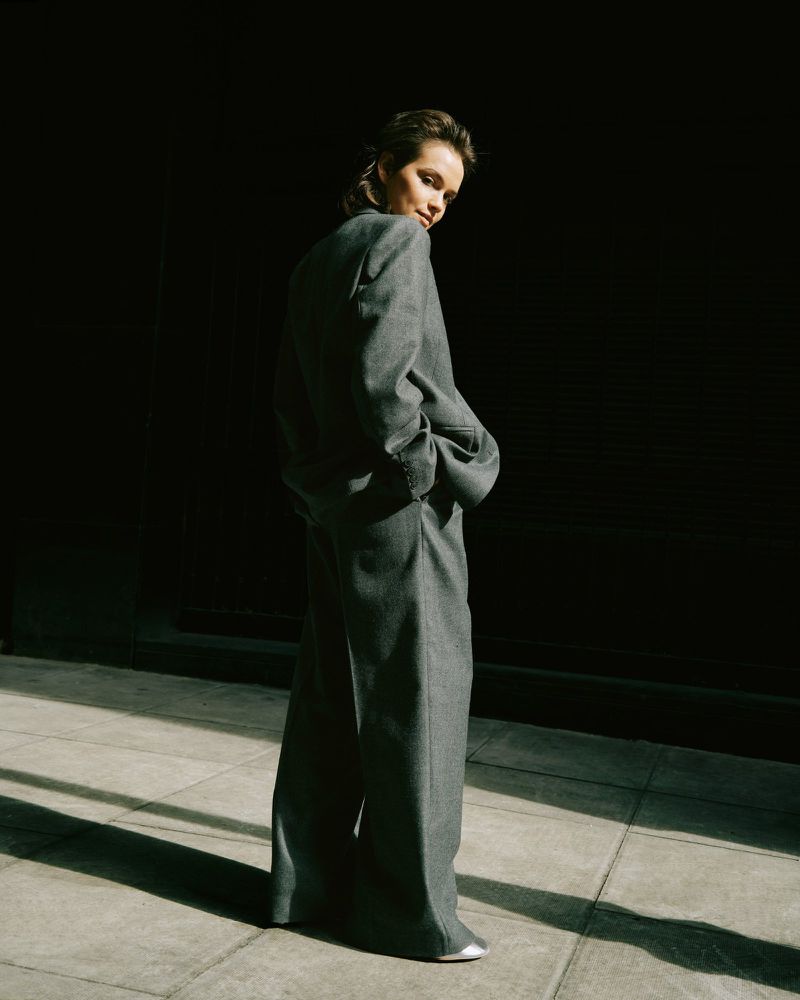 A young adult woman poses in a gray suit in front of a dark wall with natural light casting shadows across the stone floor.