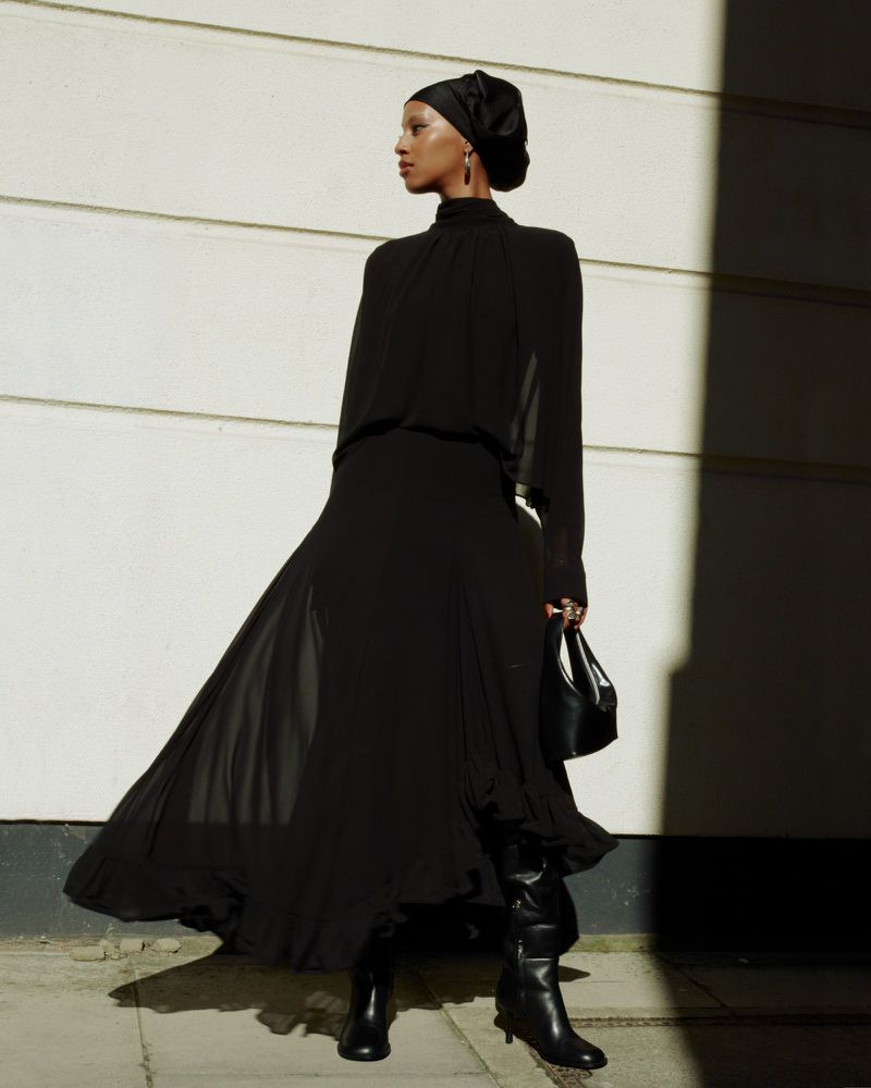 A woman in a long black dress and head covering poses against a white wall with shadows, accessorized with black boots and handbag.