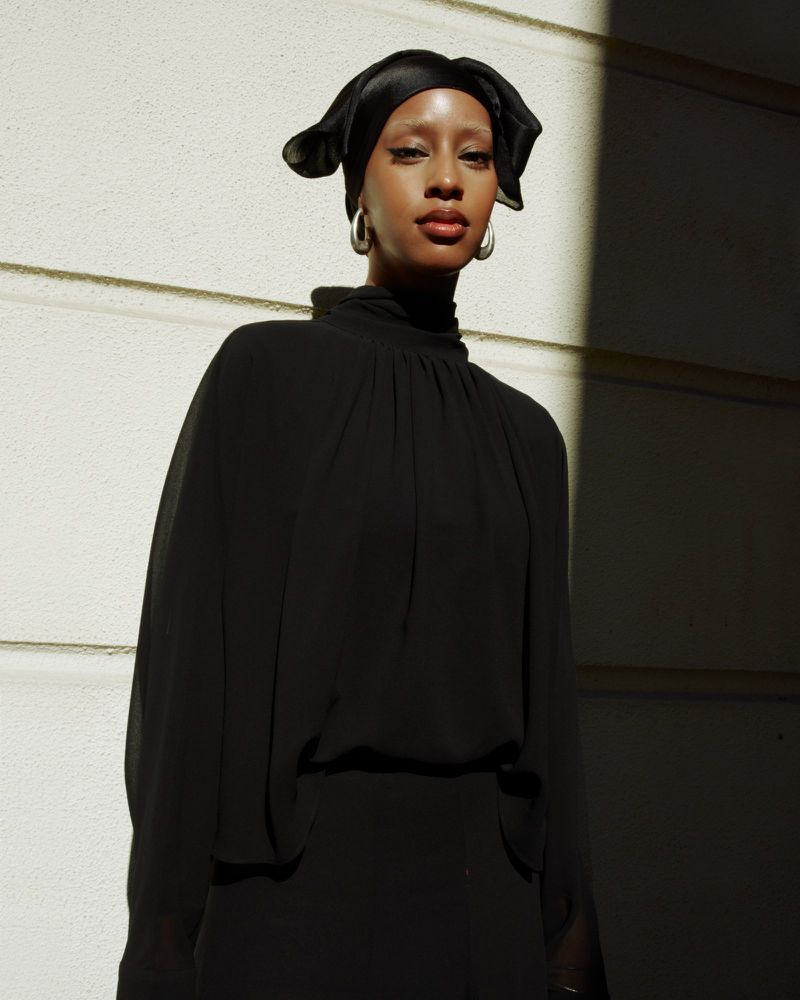 Elegant portrait of an adult woman in a black high-necked top and headscarf, standing indoors near a sunlit wall. Fashion editorial.