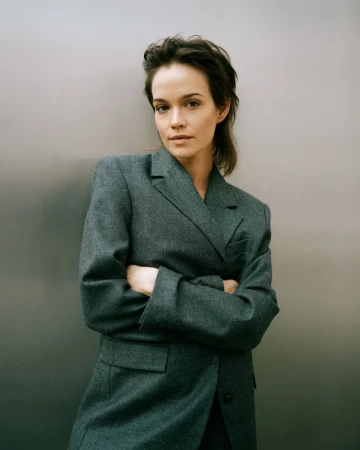 Brunette woman in a grey blazer stands with crossed arms against a grey background in a medium shot capturing her confident pose.