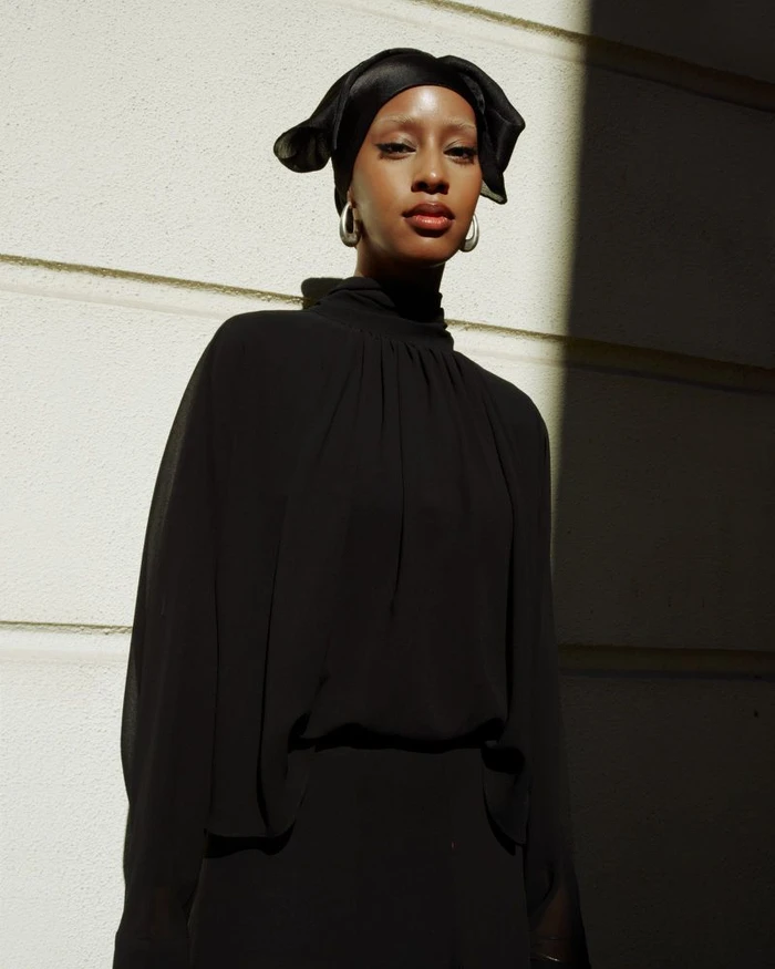 Model wearing a black top and matching head covering with silver hoop earrings against an exterior wall with hard sunlight.