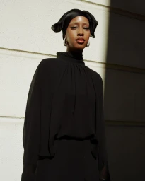 Model wearing a black top and matching head covering with silver hoop earrings against an exterior wall with hard sunlight.