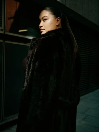 Woman wearing a dark faux fur coat looking back over her shoulder in a dimly lit urban setting at night