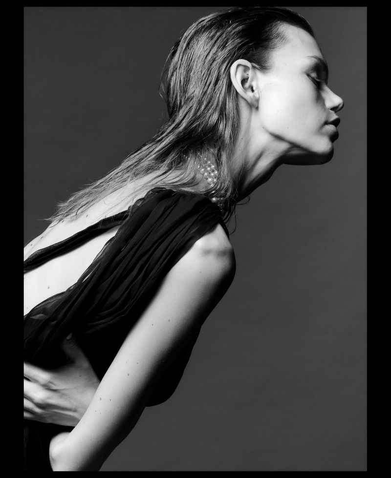 Monochrome studio portrait of model Daphne Maas with wet hair and a pearl necklace against a grey background.