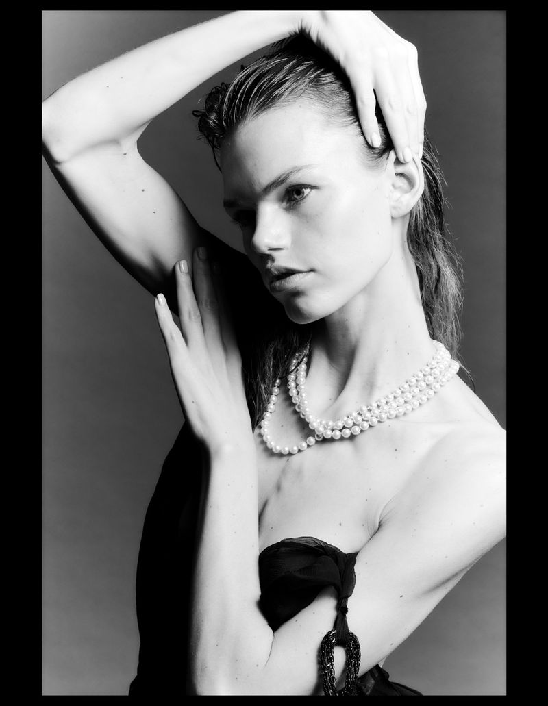 Black and white portrait of a woman with wet hair and pearl necklace, holding her hands up, against a grey background.