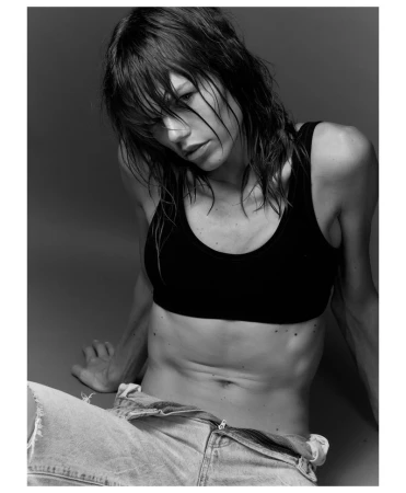 Model Daphne Maas with wet dark hair wearing a black crop top and denim shorts against a grey studio backdrop.