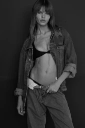 Model Daphne Maas wearing an unbuttoned denim jacket and jeans against a dark background in a monochrome studio portrait.