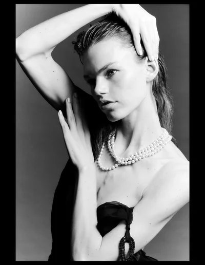 Monochrome studio portrait of model Daphne Maas wearing a multi-strand pearl necklace and posing with her hands raised.