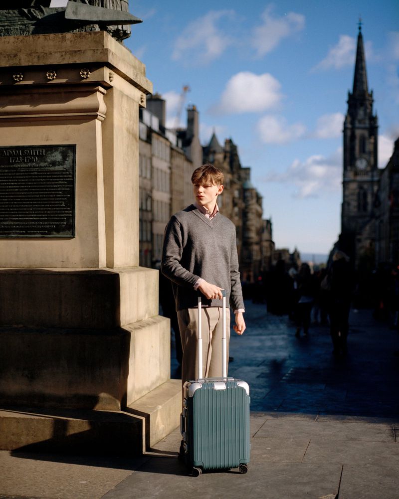 A young adult male stands on a city street next to a stone monument, holding a rolling suitcase in an urban travel scene.