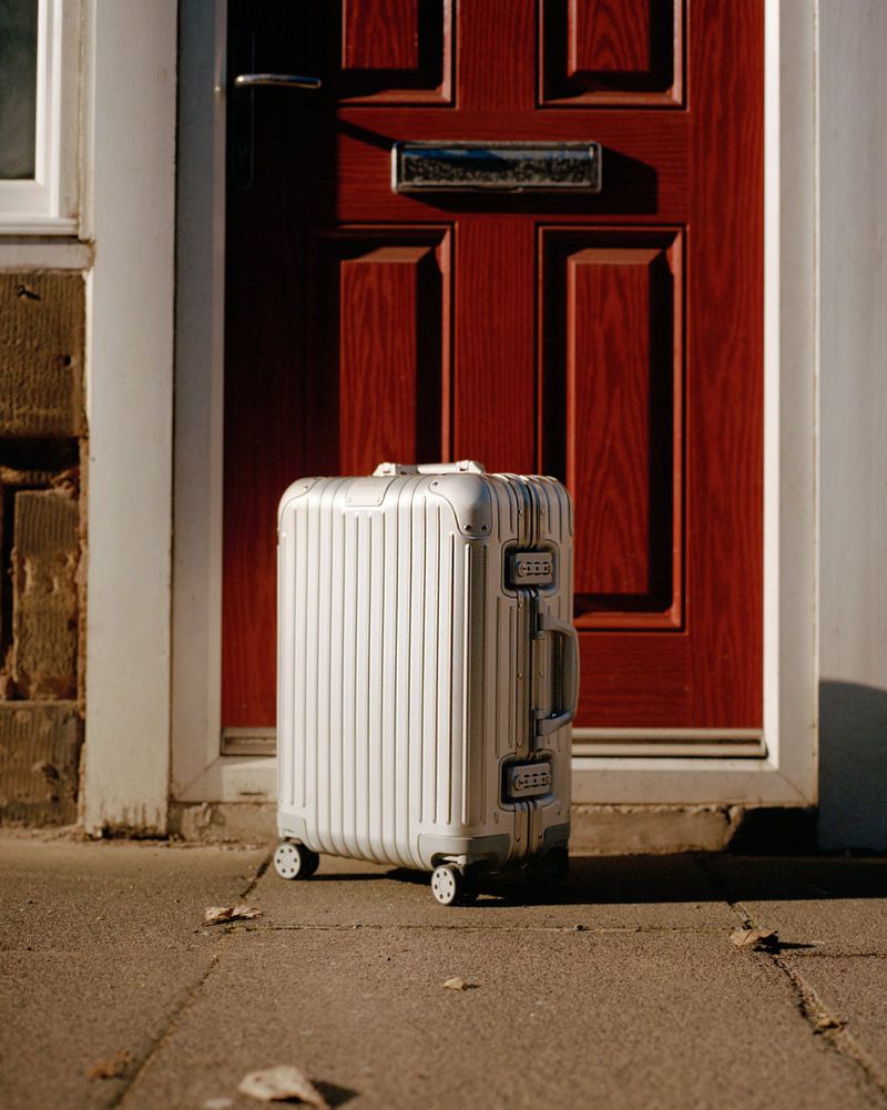 Silver carry-on suitcase on wheels sits on a sidewalk in front of a red paneled door with gray trim on a sunny day.