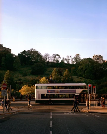 Urban scene of a double-decker bus and pedestrians crossing a city street against a backdrop of green hills and a castle.