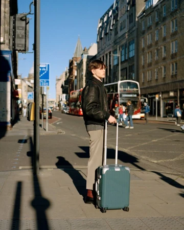A stylish young man with brown hair stands on a city street with a blue suitcase, wearing a black jacket, tan pants, and red shoes.