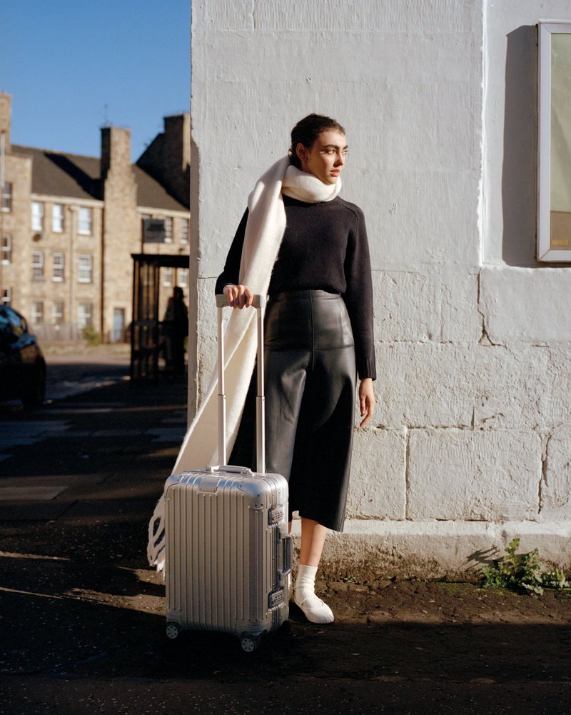 A woman stands outside next to a silver suitcase in natural lighting. She wears a dark sweater, culottes, socks, and a cream scarf.