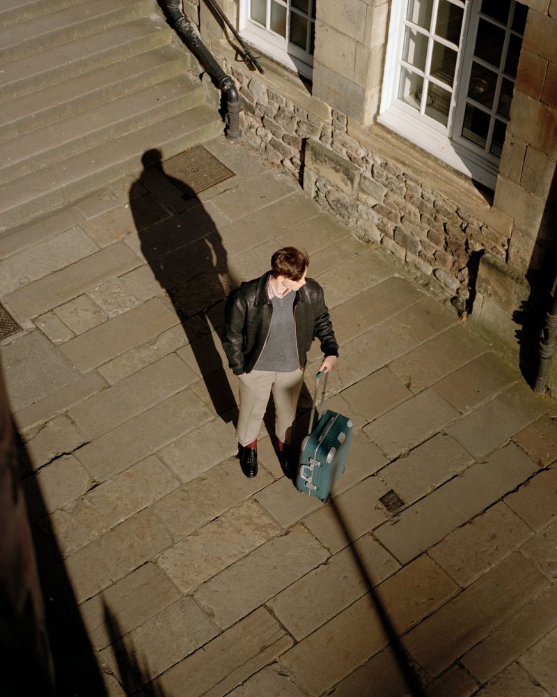 High angle shot of a man standing on a stone street next to stone steps, pulling a wheeled suitcase, with the shadow cast to the left.