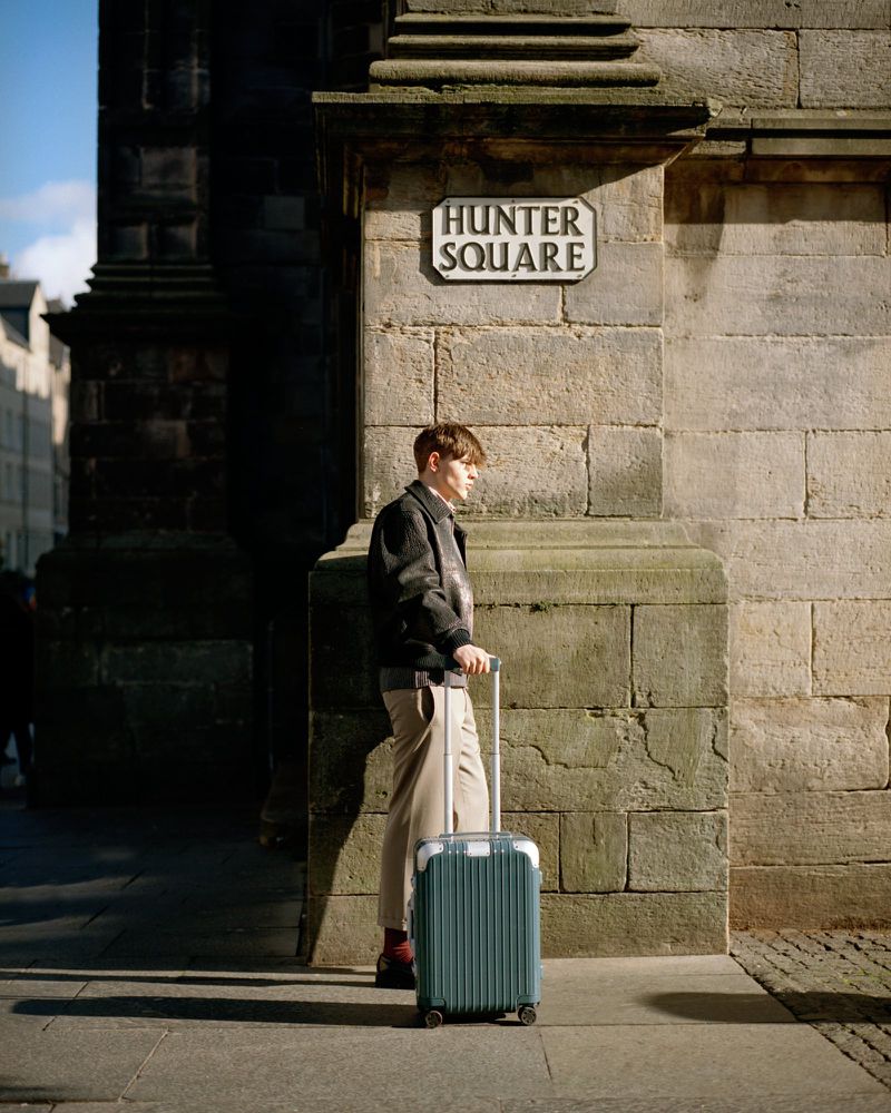 A young adult male stands on a city sidewalk with a blue suitcase near a building corner under a Hunter Square street sign.