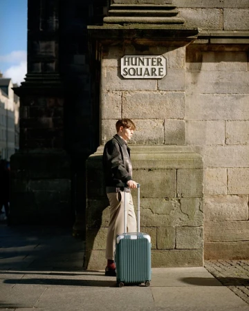 A young adult male stands on a city sidewalk with a blue suitcase near a building corner under a Hunter Square street sign.